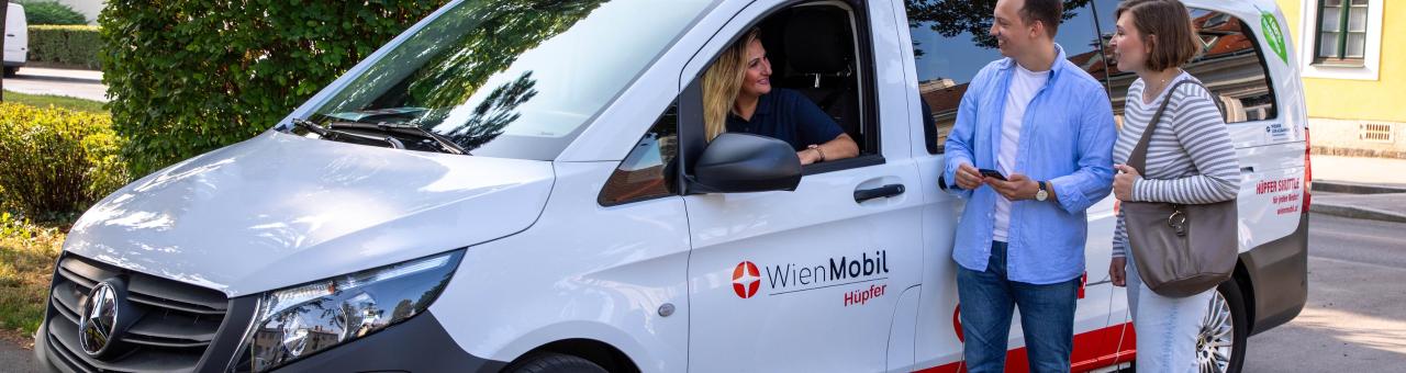 eine Frau sitzt in einem Kastenwagen mit der Aufschrift Wien Mobil Hüpfer sie lächelt zwei Personen zu die neben dem Fahrzeug stehen