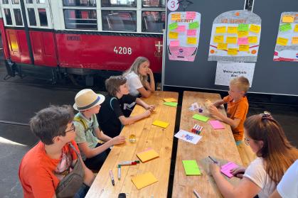 An einem Tisch sitzen mehrere Kinder, die auf buntes Papier zeichnen. Im Hintergrund sind eine alte Straßenbahn und eine Pinnwand.