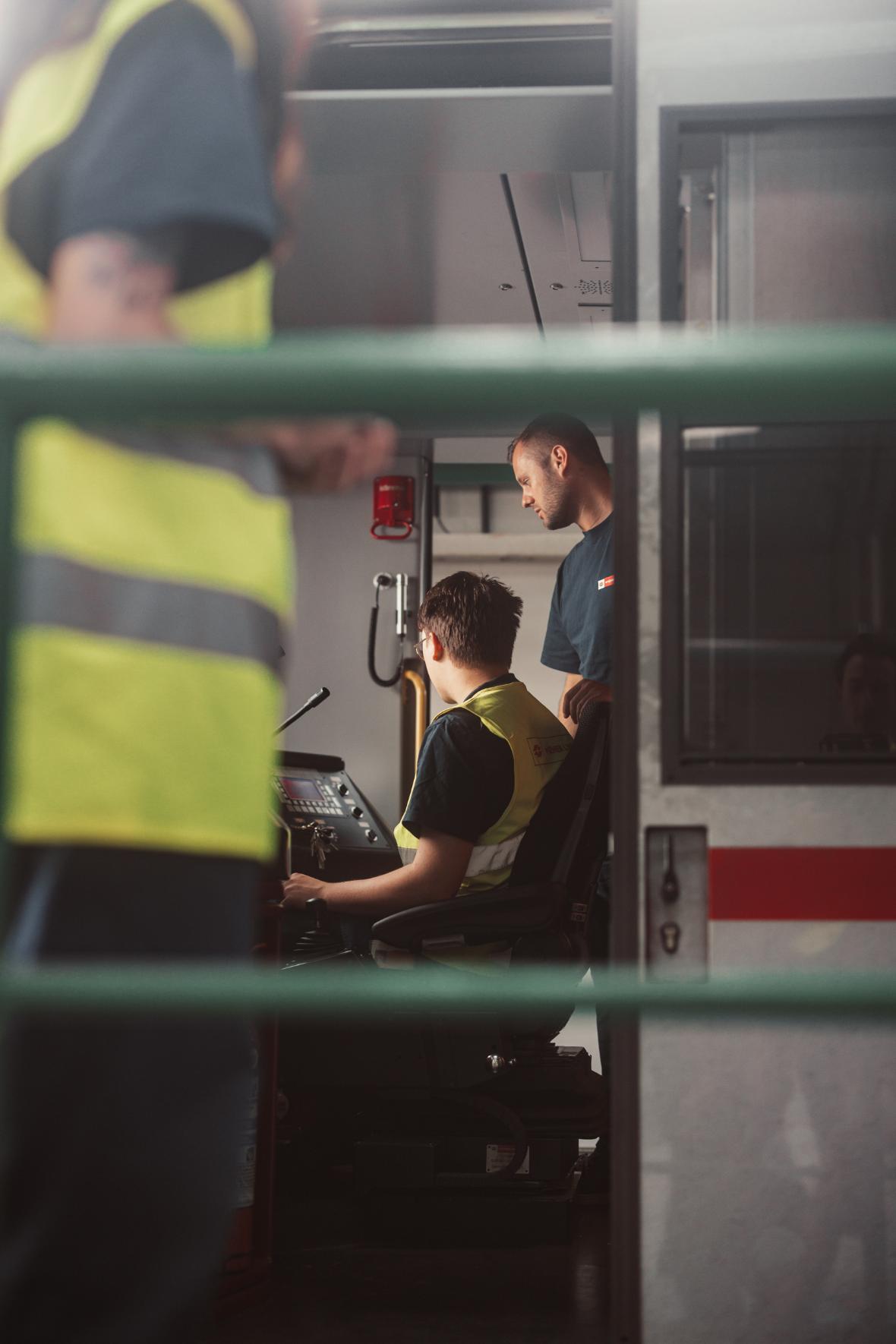 Young persons in high-visibility waistcoats are doing maintenance work in the driver's cab of an underground train
