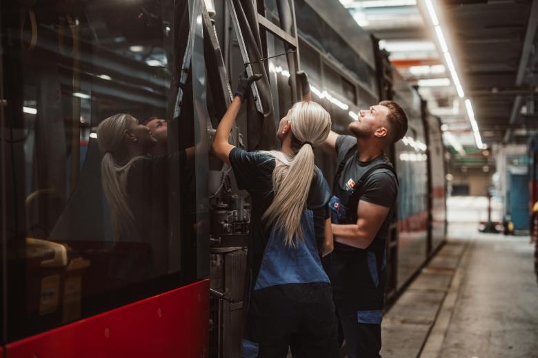 eine junge Frau mit langen blonden Haaren und ein junger Mann in Wiener Linien Arbeitskleidung montieren ein Teil von einer Straßenbahn
