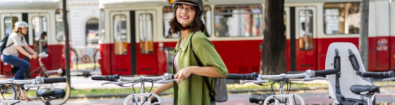 junge Frau steht mit Fahrradhelm bei der WienMobil Rad Station. Im Hintergrund fährt eine Straßenbahn vorbei.